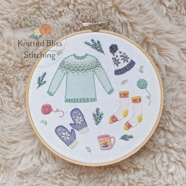 Embroidery hoop with knitted-themed design on a fluffy beige surface, featuring 'Knitted Bliss Stitching' brand.