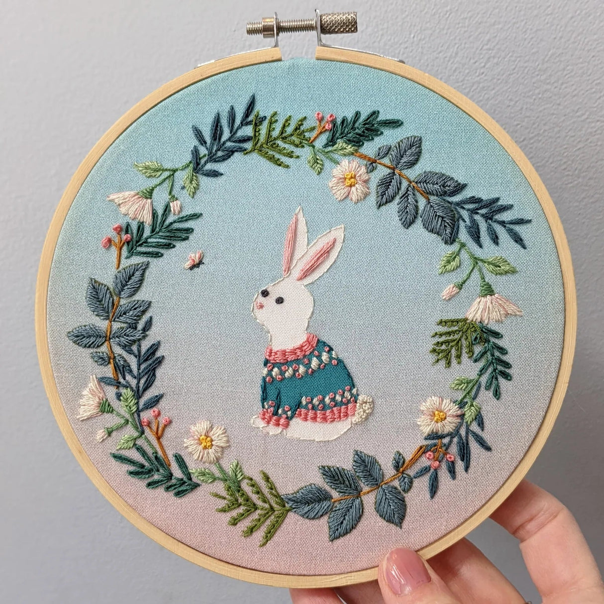Embroidery hoop with a rabbit and floral design held against a gray background
