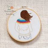 Embroidery hoop with a person wearing a rainbow sweater on a textured fabric background, featuring 'Knitted Bliss Stitching' logo.