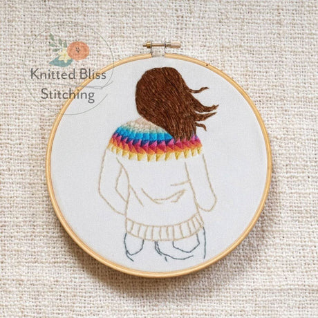 Embroidery hoop with a person wearing a rainbow sweater on a textured fabric background, featuring 'Knitted Bliss Stitching' logo.