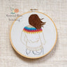 Embroidery hoop with a person wearing a rainbow sweater on a textured fabric background, featuring 'Knitted Bliss Stitching' logo.
