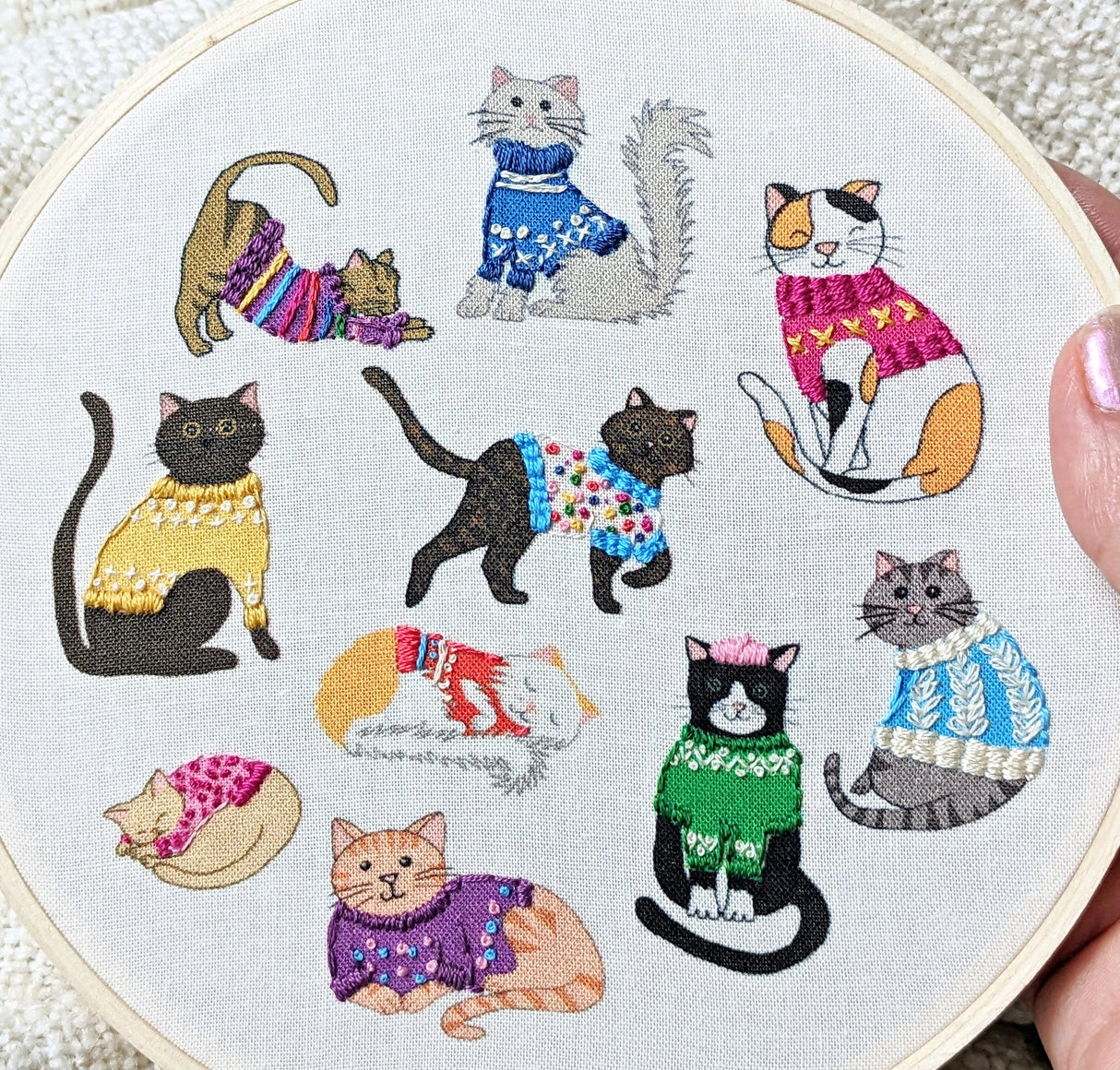 Embroidery hoop with embroidered cats wearing colorful sweaters on a white background.